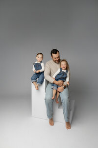 Father sitting on white studio cubes holding his two young children, minimalist gray backdrop Yucaipa studio family portrait by Kaitlyn Dawn Photography.