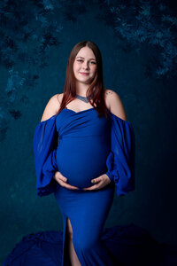 Radiant mother in a royal blue off-the-shoulder gown stands in front of a rich blue backdrop, her hands gently resting on her belly.