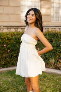High school senior girl in a white dress standing outdoors near a garden during golden hour, senior portrait by Kaitlyn Dawn Photography.
