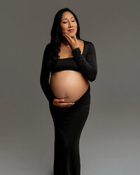 Pregnant woman in a black studio maternity dress gently holding her baby bump during a maternity portrait session by Kaitlyn Dawn Photography.