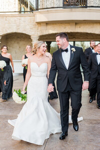 Bride and groom smile at each other, walking hand in hand followed by their wedding party