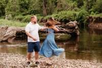 Husband and wife dancing on a river bank.  Wife is twirling in towards her husband wearing a light blue dress.  Photo taken by Philadelphia family photographer, Kristi
