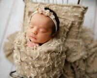Sleeping newborn baby girl wearing a floral headband and wrapped in a cream swaddle on a textured neutral blanket during a professional newborn photography session by Kaitlyn Dawn Photography in Inland Empire.