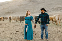 Expecting parents holding baby boots and looking at each other during a ranch maternity photography session surrounded by cattle at sunset.
