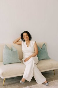 Woman wearing an all-white outfit and leaning on her elbow on a couch with blue pillows.