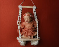 Sleeping newborn baby girl posed in a miniature swing prop with a warm red backdrop during a creative studio newborn photography session with newborn photographer Kaitlyn Dawn Photography.