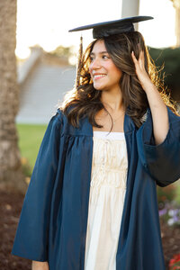 High school graduate wearing a blue cap and gown adjusting her cap in warm sunset light, senior graduation portrait by Kaitlyn Dawn Photography.
