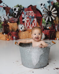Baby boy enjoying a bubble bath in a galvanized tub after his farm-themed cake smash session, with a barn backdrop and cake on his face.
Photograph by Yucaipa cake smash photographer Kaitlyn Dawn Photography.