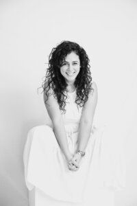A black and white picture of a woman with dark curly hair smiling widely while sitting on a white box against a white backdrop. She wears a long white dress and holds her hands together in front of her.