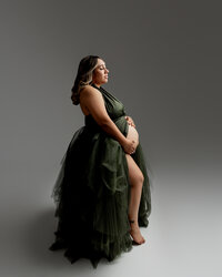 Studio maternity portrait of expecting mother wearing a dark emerald green maternity gown posing in profile with hands on baby bump photographed by Yucaipa best maternity photographer Kaitlyn Dawn Photography.