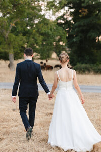 A couple, dressed in a wedding gown and suit, walk hand in hand on a grassy path at Wasing Park. They are facing away from the camera, heading toward trees and a group of grazing animals in the background, embodying the serene beauty of their special day.