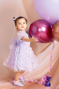 A toddler girl stands in a purple dress holding giant balloons.