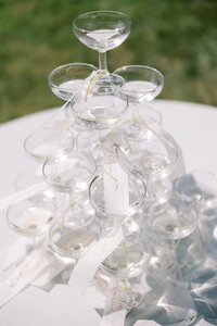 Stacked champagne coupe glasses arranged in a tower with delicate name tags, photographed outdoors as an elegant wedding reception detail in soft natural light.