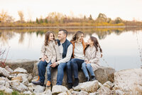 Four people sitting on rocks by a calm lake with trees in the background, sharing smiles during a Toronto family photography session.