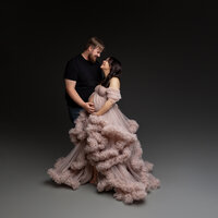 Romantic maternity studio portrait of expecting couple embracing with mother wearing a dramatic blush ruffled maternity gown and father gently holding her baby bump during maternity studio session by Kaitlyn Dawn Photography.