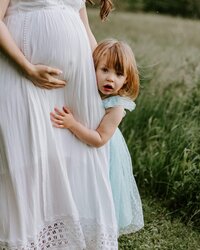 A pregnant woman in a white dress holding a little girl in a field.