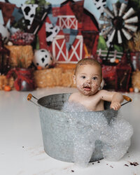 Farm-themed cake smash session ends with a cheerful bubble bath baby boy in a tin tub with a barn backdrop and frosting on his face.
Photograph by Yucaipa cake smash photographer Kaitlyn Dawn Photography.