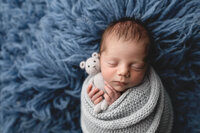 Baby boy holding a grey teddy wrapped in a matching blue-gray knitted wrap at Jennifer Brandes Photography studio.