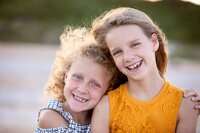 Two sisters smiling with their heads leaned together and arms wrapped around each other during a beach family portrait session. (Photographed by Nature's Child Photography)