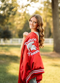 Senior portrait in downtown Magnolia Springs, Alabama — looking back at the camera with her dance team jacket draped over her shoulder.