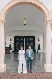 A bride and groom stand separately under an arched entryway decorated with christmas wreaths, looking in opposite directions.