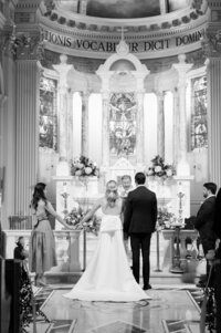 A bride and groom stand before an officiant in an ornate church, holding hands with their wedding party beside them.