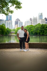 Engaged couple smiling in Piedmont Park with the city skyline in the background.