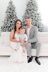 family of three sitting on a white couch with Christmas trees in the background by Miami Christmas Minis Photographer MSP