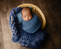 Newborn baby boy peacefully sleeping while wrapped in a soft blue swaddle and posed in a wicker basket during a studio newborn photography session by Kaitlyn Dawn Photography.