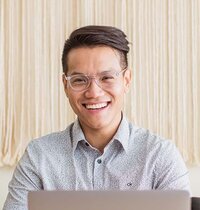 A handsome, light-skinned Asian man with stylish hair and glasses, smiles broadly from behind a laptop