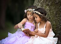 Two young girls dressed as princesses in soft lavender and blush gowns read from an vintage poetry book beneath a tree during a signature fairy adventure session in St. Johns. (Photographed by Nature's Child Photography)