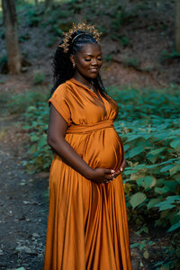 Pregnant woman in a rust orange gown and crown cradling her belly on a forest path during a Cleveland maternity photography session