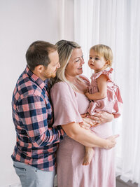 Pregnant mom is holding her little daughter while dad hugs them from behind