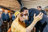 A man in a suit tenderly touches the face of a woman in a yellow outfit at a lively gathering, with onlookers in the background.