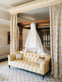 A classic white wedding gown hanging above a tufted yellow sofa at Dover Hall, illustrating the refined composition of a luxury wedding weekend.