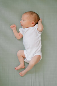 A newborn boy in a white onesie, laying on top of his green crib sheet sleeping