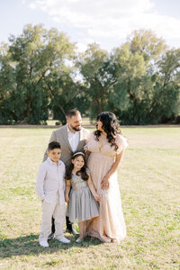Dilorio family standing together in a sunlit Las Vegas park, with parents smiling at each other and their two children posing in a natural outdoor family portrait session.