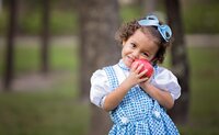 Preschool-aged girl with curly hair and a blue bow smiling while holding a red apple in a wooded St. Johns, park during a child photo session. (Photographed by Nature's Child Photography)