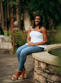 Senior portrait at Bella Sera Gardens in Loxley, Alabama — sitting on the fountain with greenery
in the foreground.