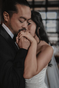 Couple de mariés enlacés, les yeux fermés, partageant un moment intime et tendre lors de leur première danse, le marié embrassant la main de la mariée, baigné de lumière naturelle et d’émotion.