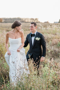The couple walks through a golden field at sunset, the bride lifting her dress slightly as they navigate the tall grass.
