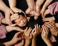 Family members’ hands surrounding and supporting a sleeping newborn baby wrapped in black during a creative studio newborn photography portrait by Yucaipa newborn photographer Kaitlyn Dawn Photography.