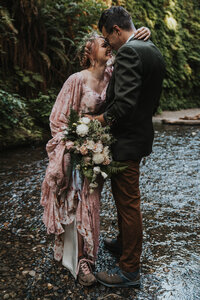 Bride wearing rose pink gown holds groom close to her both wearing hiking boots standing in shallow creek in Humboldt creek by California elopement photographer Kasey Mantiply