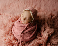 Sleeping newborn baby girl wrapped in a blush pink swaddle and wearing a bonnet while posed on a soft pink textured blanket during a newborn photography session.