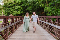 Expectant couple walking hand in hand across a wooden bridge during a Youngstown maternity photography session
