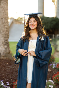 High school graduate smiling in a blue cap and gown outdoors during golden hour, senior portrait by Kaitlyn Dawn Photography.