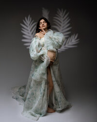 Fine art pregnancy portrait of a pregnant woman wearing a flowing green maternity gown with soft botanical light patterns photographed by Kaitlyn Dawn Photography.
