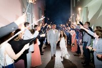 Jacksonville, FL wedding photographers photographing in St. Augustine, wedding capturing a bride and groom walking hand in hand through a nighttime sparkler exit, guests lining the walkway and celebrating around them.