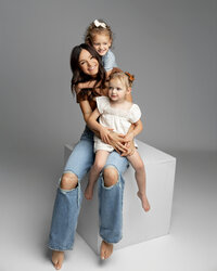 Mother sitting on white studio cube holding her two young daughters, neutral tones and soft lighting creating a modern studio family portrait by Kaitlyn Dawn Photography.