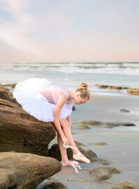 Professional photography by Nature's Child Photography: Young tween ballerina in a pink leotard and white tutu leaning forward to adjust her ballet shoe while sitting on seaside rocks during a beach dance session in Palm Coast.